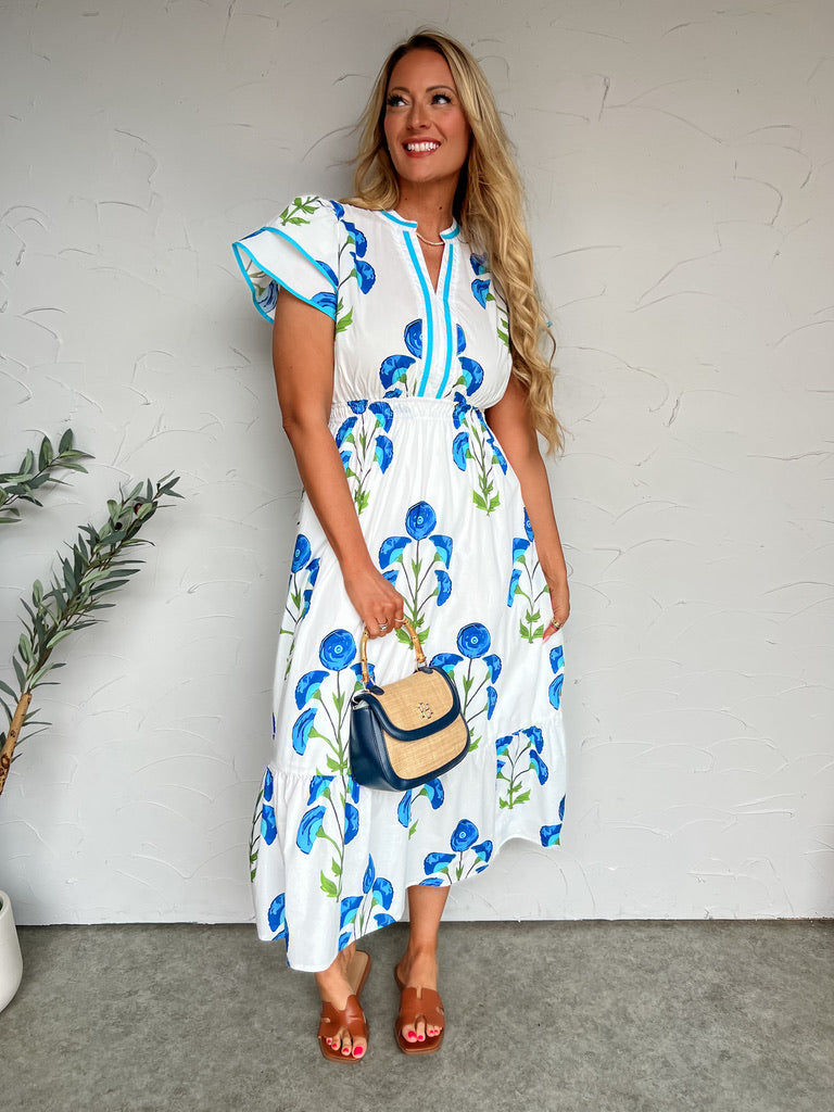 Woman wearing a white dress with blue floral patterns, holding a small handbag against a plain background.