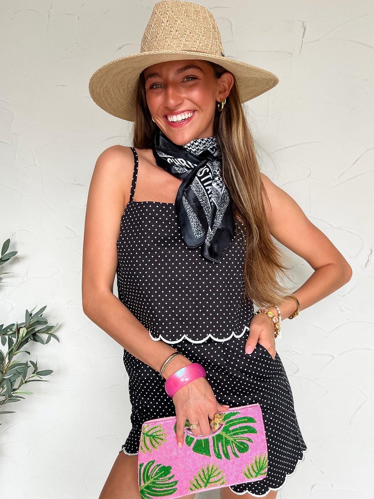 Woman wearing a black polka dot romper, straw hat, and pink clutch with green palm leaf design.