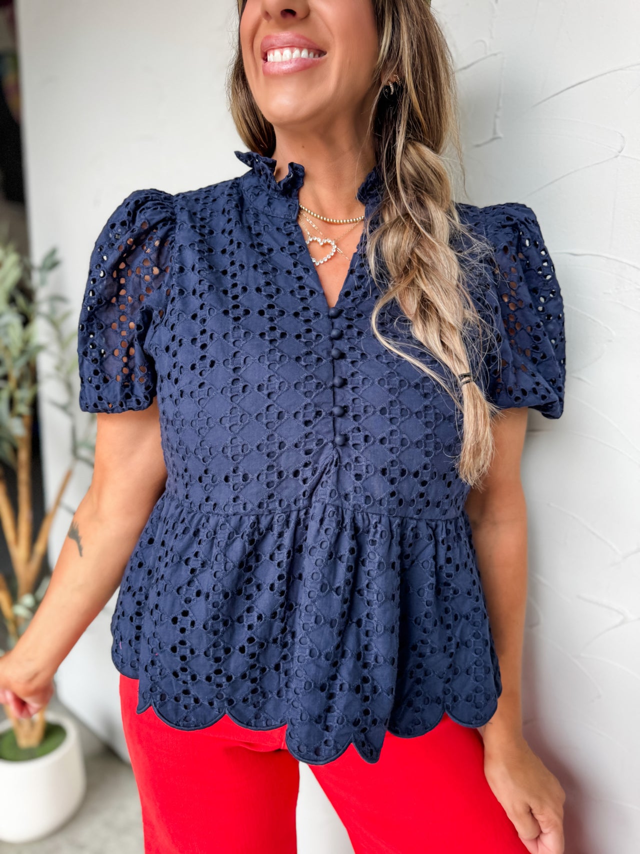 A close-up shot of a woman smiling and leaning against a textured off-white wall, wearing a navy blue short-sleeve top crafted from a 100% cotton eyelet fabrication. The top features an intricate all-over geometric eyelet design, a ruffled stand collar, an open V-neckline with a functional loop-button placket down the bodice, and puffed shoulders. A gathered seam forms a flared peplum waist that finishes with a delicate scalloped bottom hem. She is styled with bright red pants and layered gold necklaces, in