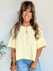 Woman wearing a yellow cable knit shirt sleeve sweater top and blue jeans standing against a white wall with a plant to the left