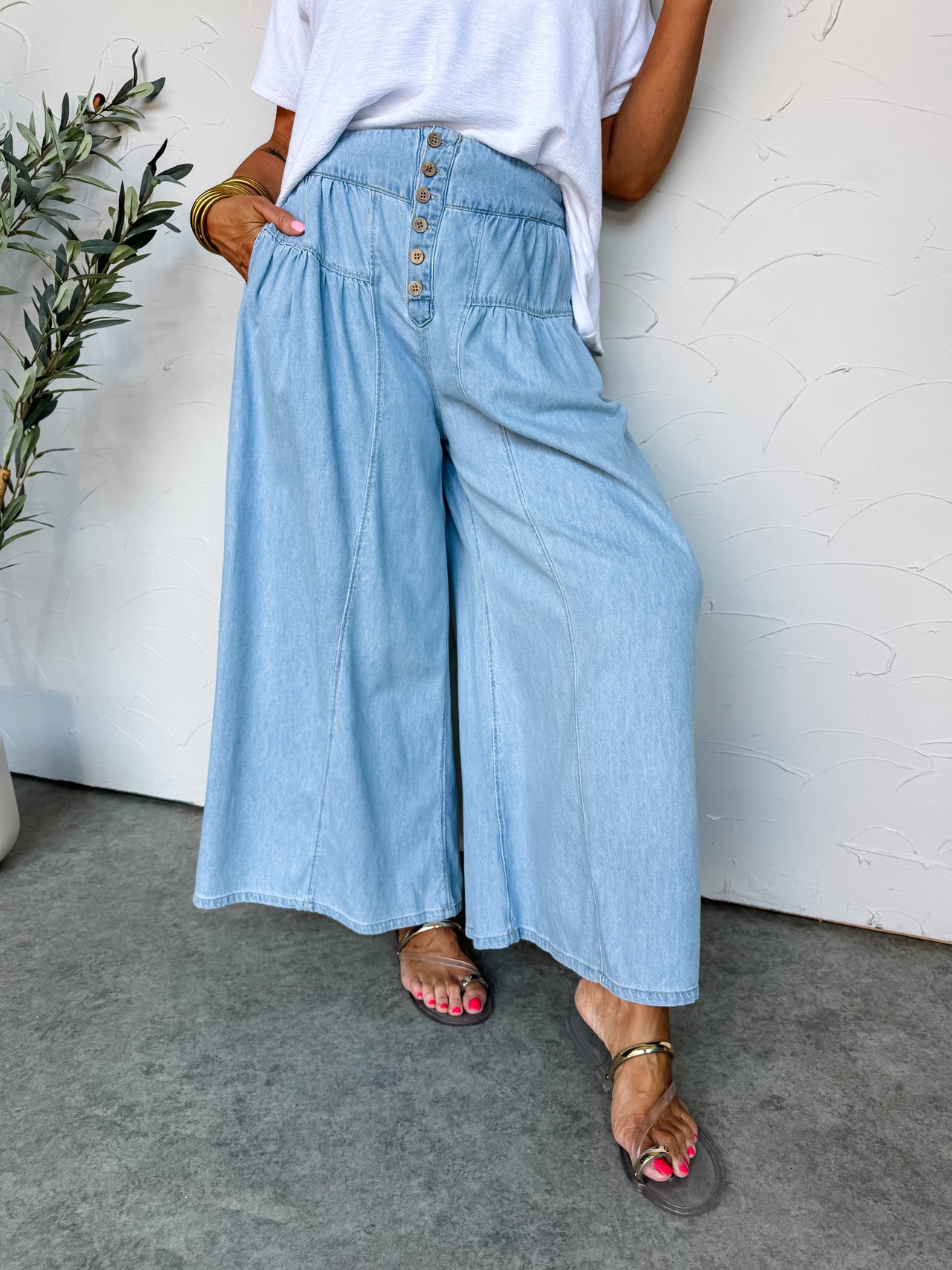 A close-up and lower-body view of a person standing against an off-white wall, wearing light-wash wide-leg denim pants made from 100% cotton. The high-waisted pants feature a fitted waistband with an exposed six-button fly, a curved tiered hip yoke with soft gathered pleating, and a relaxed, extra-wide leg silhouette. They are styled with a tucked-in white short-sleeve top and metallic gold flat sandals.