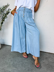 A close-up and lower-body view of a person standing against an off-white wall, wearing light-wash wide-leg denim pants made from 100% cotton. The high-waisted pants feature a fitted waistband with an exposed six-button fly, a curved tiered hip yoke with soft gathered pleating, and a relaxed, extra-wide leg silhouette. They are styled with a tucked-in white short-sleeve top and metallic gold flat sandals.