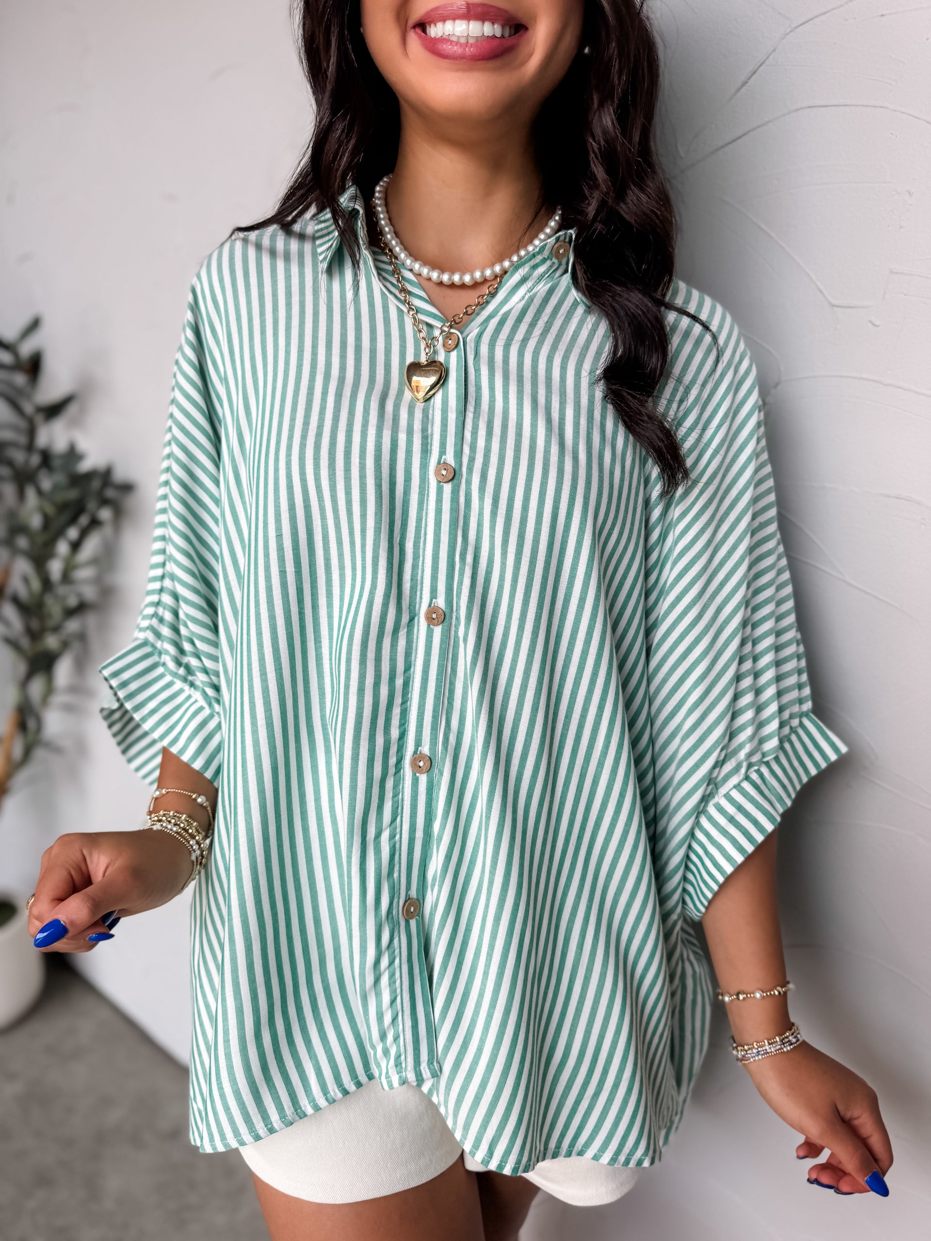 A close-up photograph from the chin down of a smiling brunette woman posing against a textured white wall. She is wearing an oversized button-down shirt made from a lightweight 100% rayon fabric, featuring a green and white vertical stripe pattern, dropped shoulders, and short cuffed sleeves.