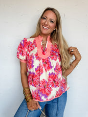 A smiling blonde woman leaning against a textured off-white wall, wearing a patterned short-sleeve top. The blouse is crafted from a 100% cotton fabric featuring an all-over bold floral print in shades of coral pink, purple, and cream. It is designed with a solid coral contrast-trimmed mandarin collar that transitions into a split V-neckline, along with coordinating coral cuffs finishing the short puffed sleeves and a matching coral trim along the bottom hem. She has the top styled with medium-wash blue den