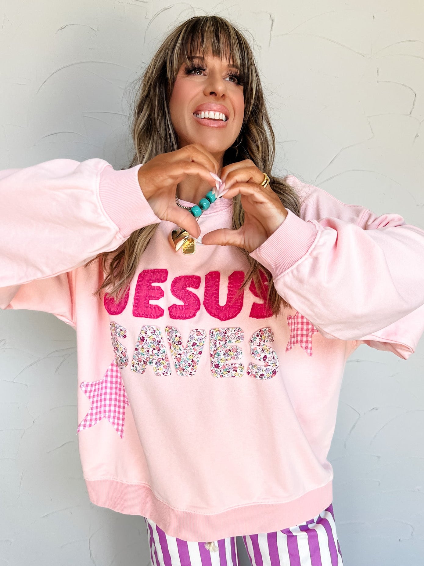 Person wearing a pink embroidered sweatshirt with 'Jesus Saves' text, making a heart shape with their hands against a light background.