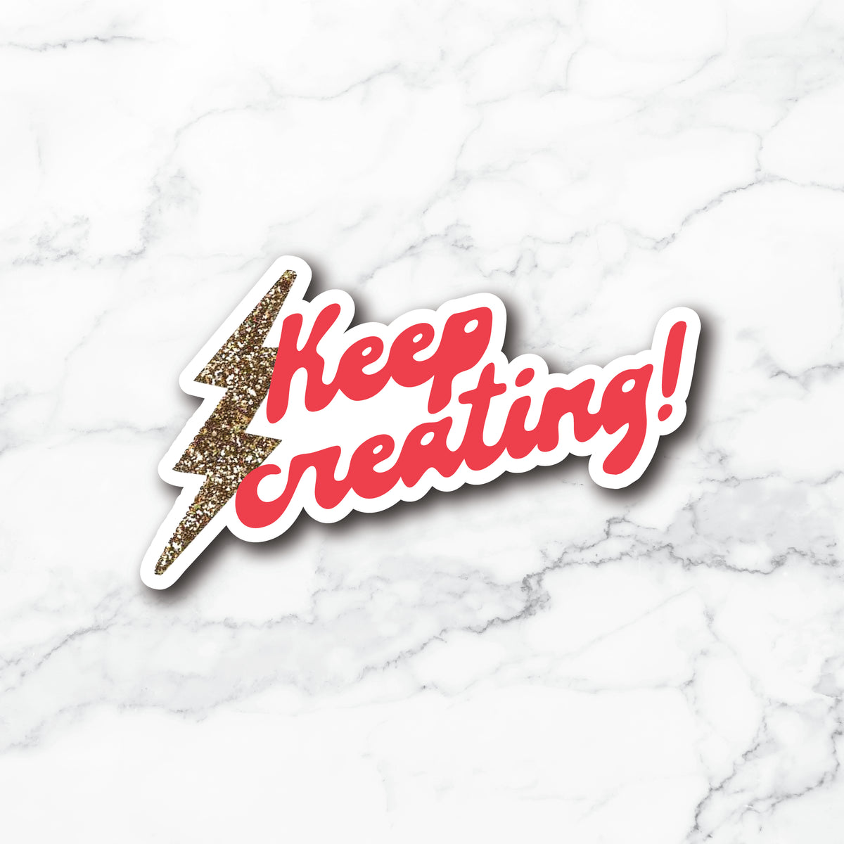 A die-cut sticker with the phrase 'Keep creating!' in red script, a glittery gold lightning bolt, and a white outline, displayed on a white marble background.