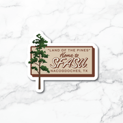 A die-cut sticker featuring a pine tree and the text “Land of the Pines Home to SFASU. Nacogdoches, TX” on a white marble background.