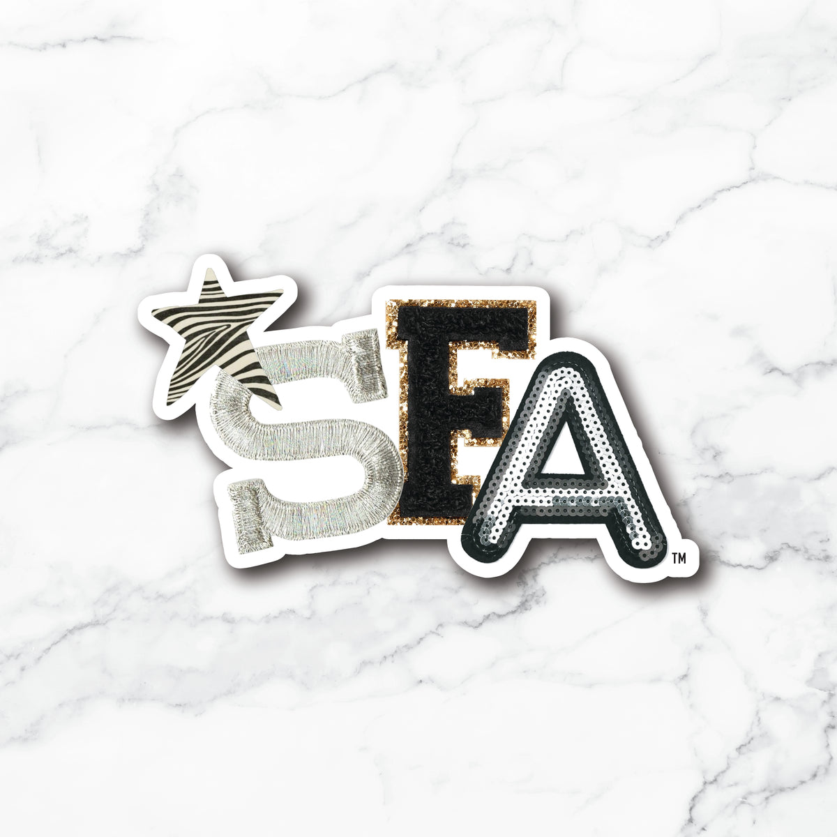 A die-cut sticker featuring the letters 'SEA' in a textured, metallic font on a white marble background. The 'S' is silver with a zebra-stripe star above it, the 'E' is black with gold glitter trim, and the 'A' is black with silver glitter trim.