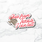 A die-cut vinyl sticker featuring the phrase 'She's from Texas, I can tell' in red script over a Texas-shaped outline filled with a brown and black zebra print, shown on a white marble background.
