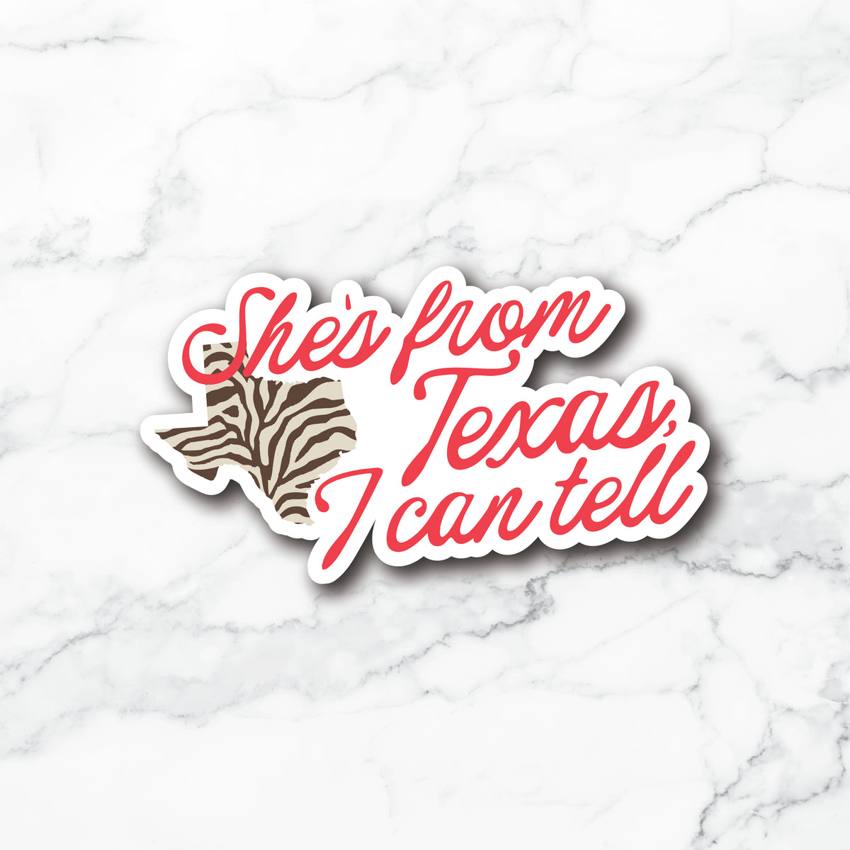 A die-cut vinyl sticker featuring the phrase 'She's from Texas, I can tell' in red script over a Texas-shaped outline filled with a brown and black zebra print, shown on a white marble background.